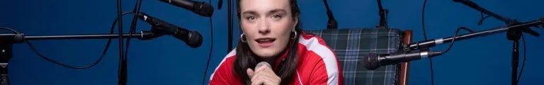Olivia Caw sitting in a plaid armchair holding a microphone against a blue background surrounded by multiple microphone stands for the Edinburgh Fringe theatre production Common Tongue.