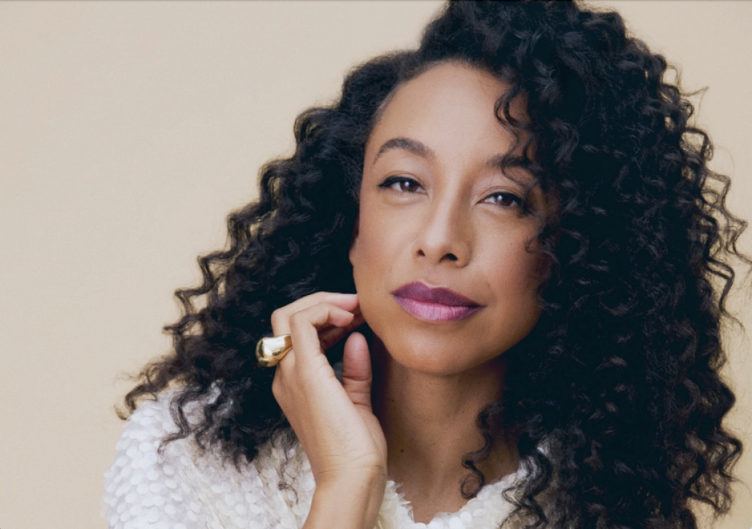 Corinne Bailey Rae with voluminous dark curly hair, wearing a white textured top and a gold ring, resting her hand near her face.