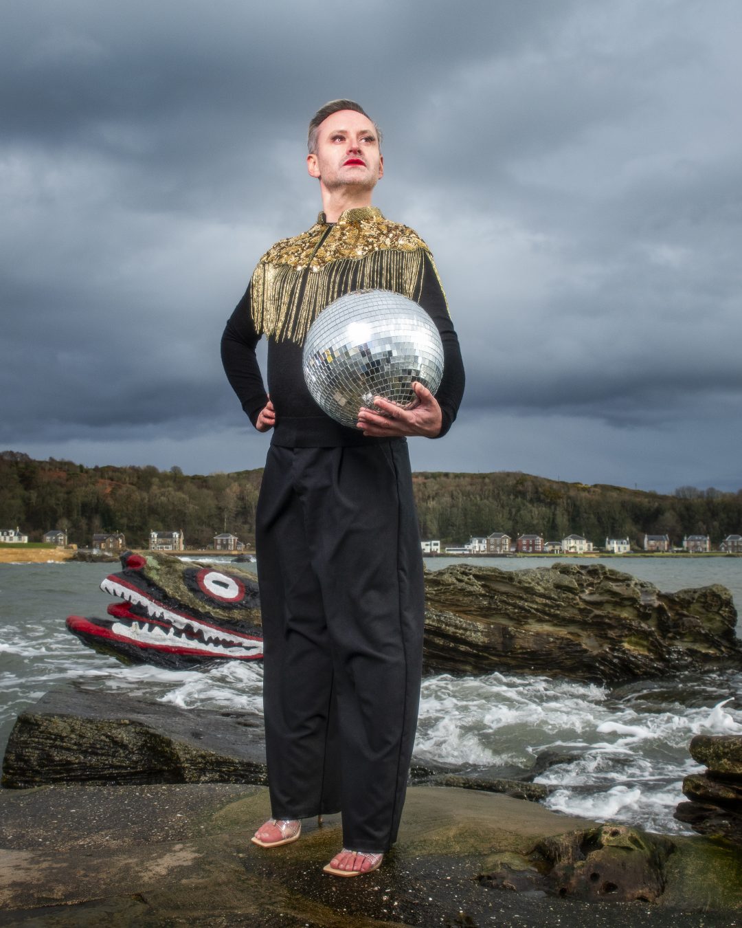 A performer holding a disco ball while standing on a coastal rock formation near the painted Crocodile Rock for Edinburgh Fringe theatre.