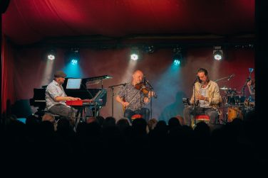 A live band performing on a stage under red lighting, featuring a keyboardist, a violinist, and a flautist in front of an audience.