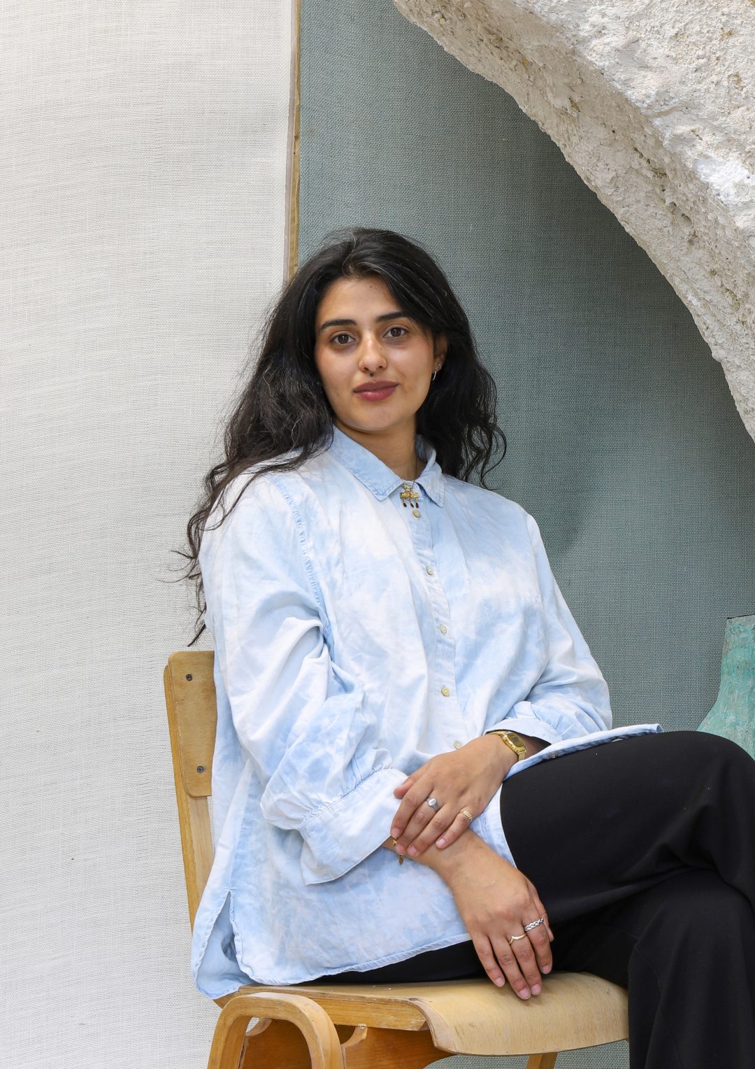 A portrait of artist Emii Alrai sitting on a wooden chair, wearing a light blue patterned shirt and black trousers, positioned in front of a textured light-coloured backdrop.