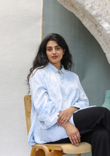 A portrait of artist Emii Alrai sitting on a wooden chair, wearing a light blue patterned shirt and black trousers, positioned in front of a textured light-coloured backdrop.