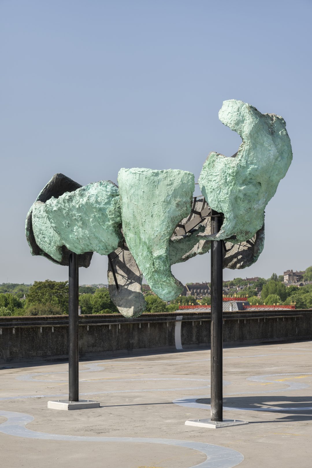 A sculptural installation by Emii Alrai titled 'Deja Ve' at Bold Tendencies. The sculpture features textured, verdigris-coloured abstract forms supported by two black pillars against a clear blue sky, situated on an outdoor rooftop level.