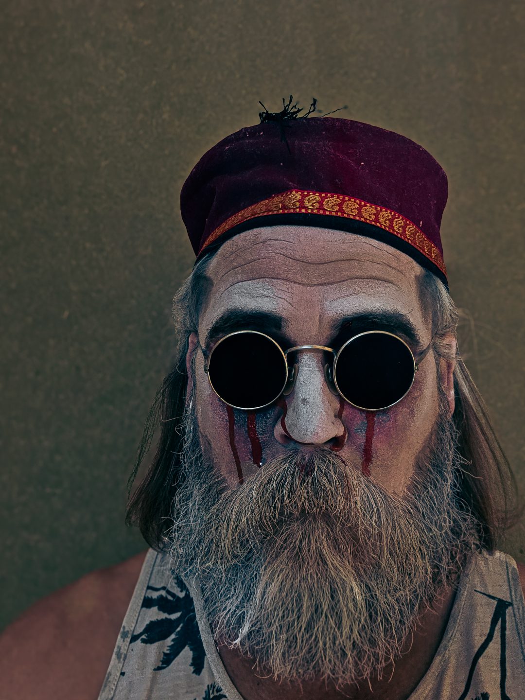 A close-up of a theatrical performer in character makeup, round sunglasses, and a red cap for Endgame.
