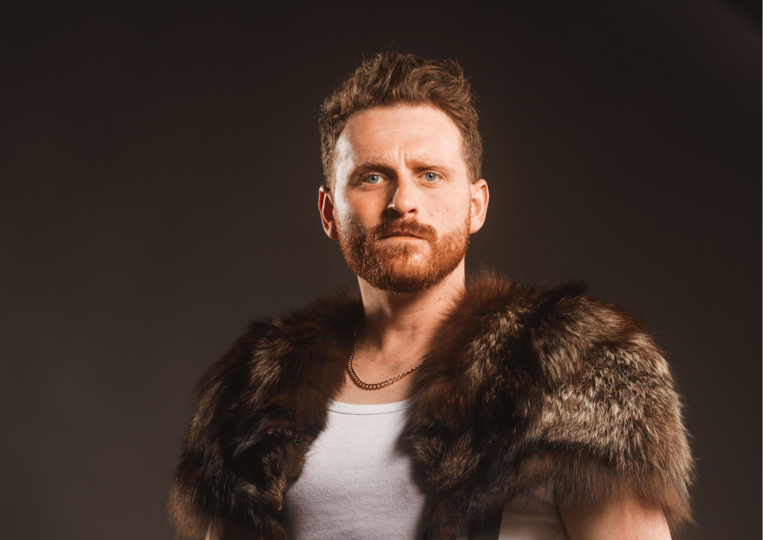 A rugged portrait of a male actor wearing a fur stole and white tank top for Heracles: Of Men and Beasts.