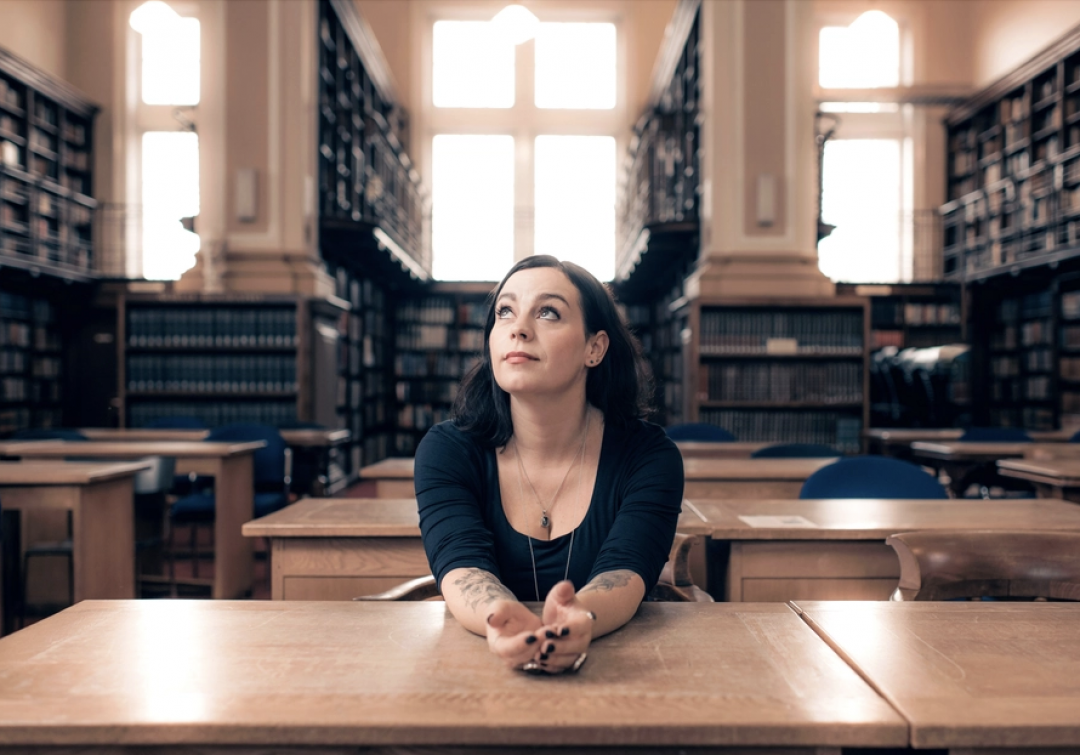 Jenni Fagan sitting at a wooden desk with her hands clasped, looking upward in a large library or archive room with tall bookshelves.