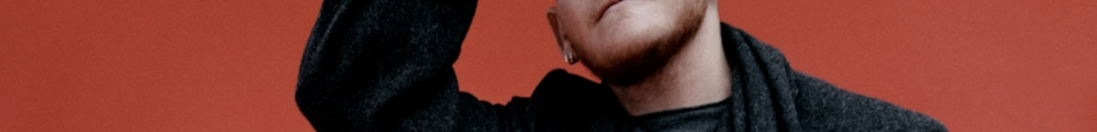 Kae Tempest wearing a grey coat and scarf against a red background, with their hand raised to their head.
