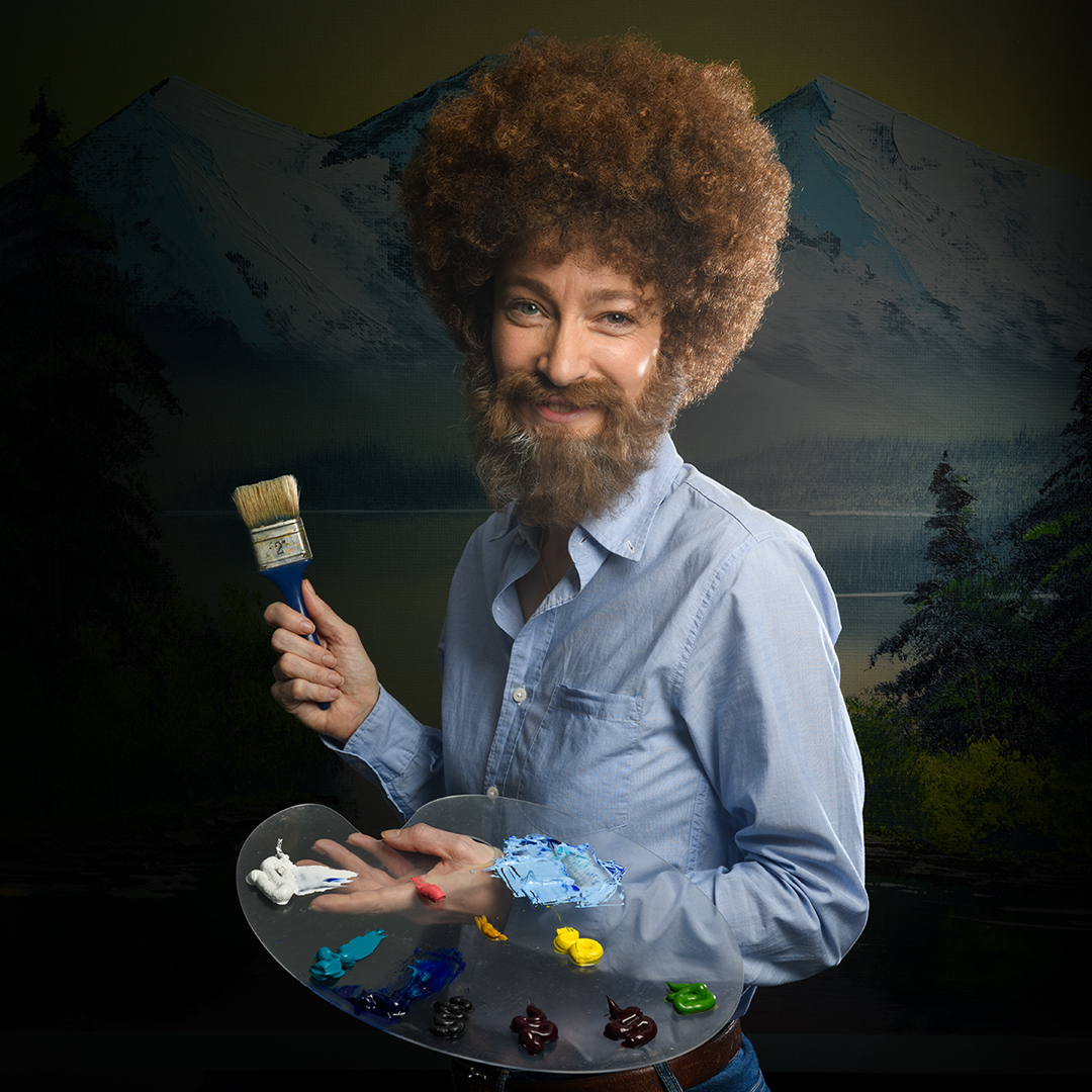 Sarah-Louise Young in costume as Bob Ross, featuring a curly afro wig and beard, holding a paintbrush against a landscape painting background.