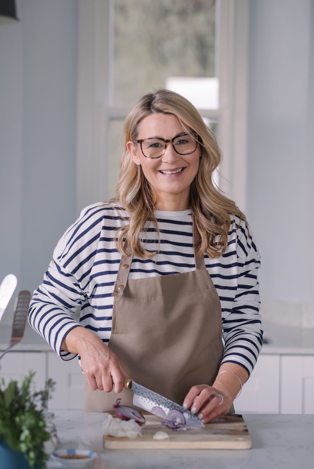 Chef Sarah Rankin smiling at the camera while slicing a red onion on a wooden board in a kitchen.