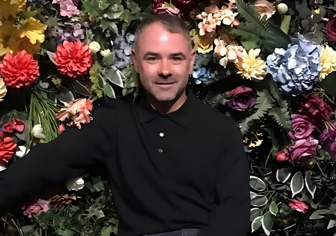 Steve Mason wearing a black polo shirt, positioned in front of a dense floral wall backdrop filled with various colorful flowers and leaves.