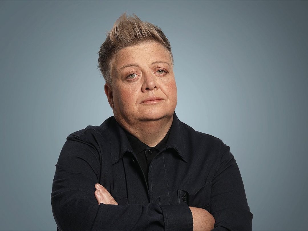 Comedian Susie McCabe posing with arms crossed against a muted grey-blue studio background