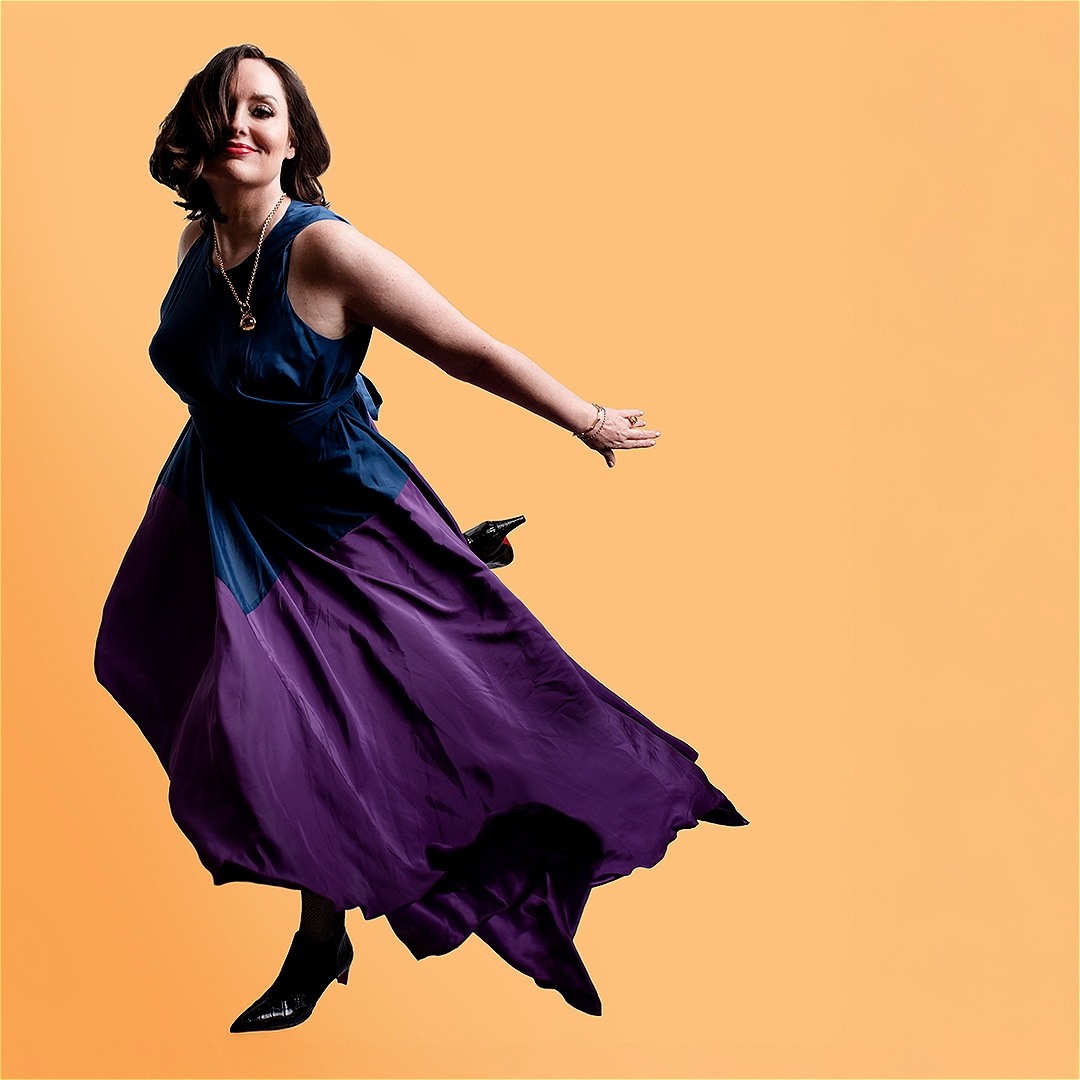 Deborah Frances-White posing dynamically in a blue and purple dress against a solid yellow background for The Guilty Feminist.