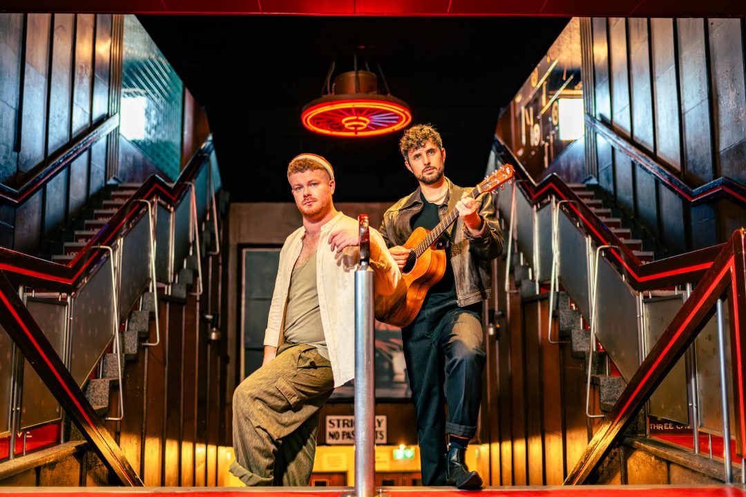 Two male musicians posing on a grand staircase with an acoustic guitar for the Edinburgh Fringe theatre production The Singer.
