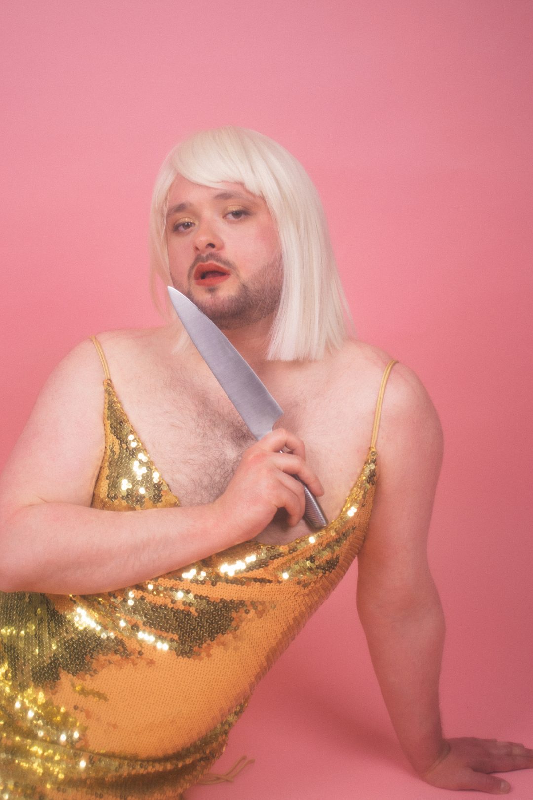 A close-up portrait of Craig Manson in a blonde bob wig and gold sequined dress, holding a large knife near his chest against a pink background.