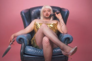 Craig Manson, in a blonde bob wig and gold sequined dress, lounging confidently in a blue leather armchair, holding a knife in one hand and a microphone in the other.
