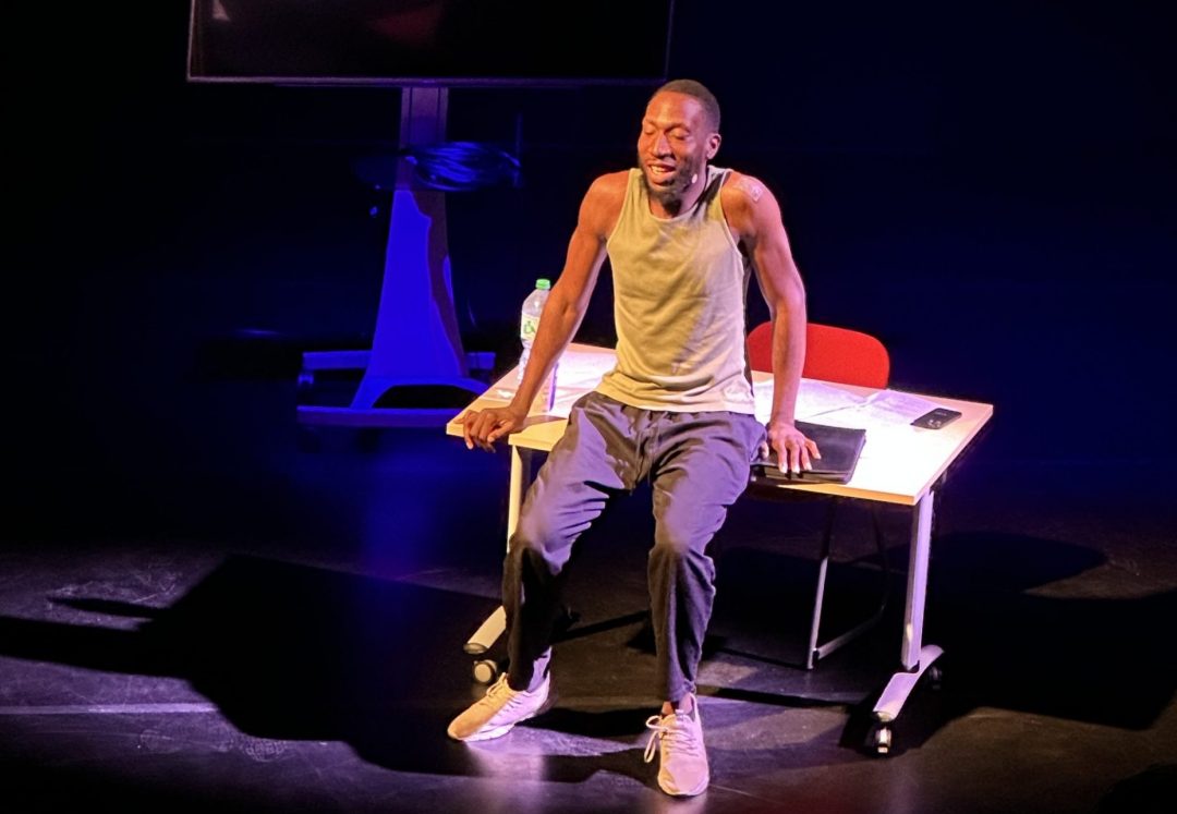 Louis Utieyin performing on stage in a tank top and trousers, sitting on a desk under a spotlight for Diary of a DL Trade.