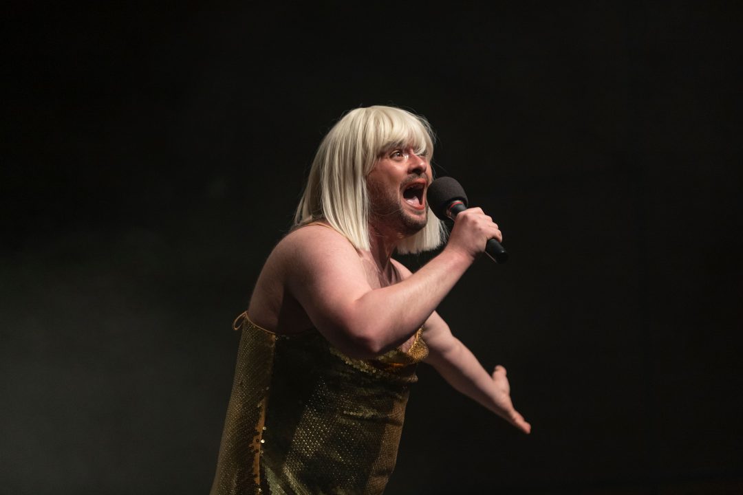 Craig Manson on stage, wearing a blonde wig and gold sequined dress, singing passionately into a microphone with arms outstretched.