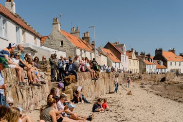 Spectators sitting along a stone wall watching a coastal event on a sunny day in a Scottish fishing village, enjoying one of the best Edinburgh Fringe alternatives