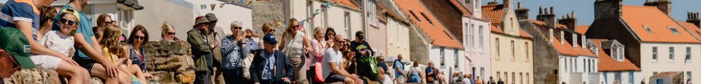 Spectators sitting along a stone wall watching a coastal event on a sunny day in a Scottish fishing village, enjoying one of the best Edinburgh Fringe alternatives