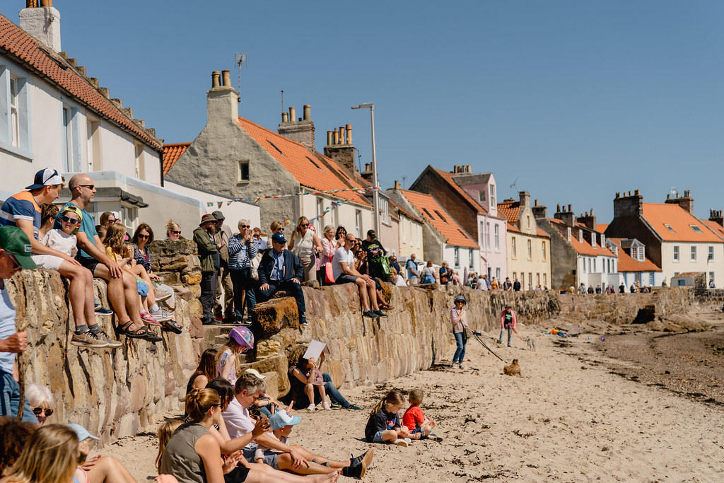 Spectators sitting along a stone wall watching a coastal event on a sunny day in a Scottish fishing village, enjoying one of the best Edinburgh Fringe alternatives