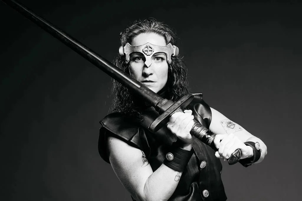 A black-and-white promotional photo of an actor in a leather costume wielding a sword for Macbeth.