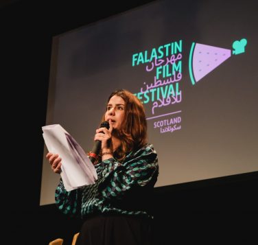 Bayan Haddah reading from notes at a microphone during the Falastin Film Festival 2024 final event, with the festival logo projected behind her.