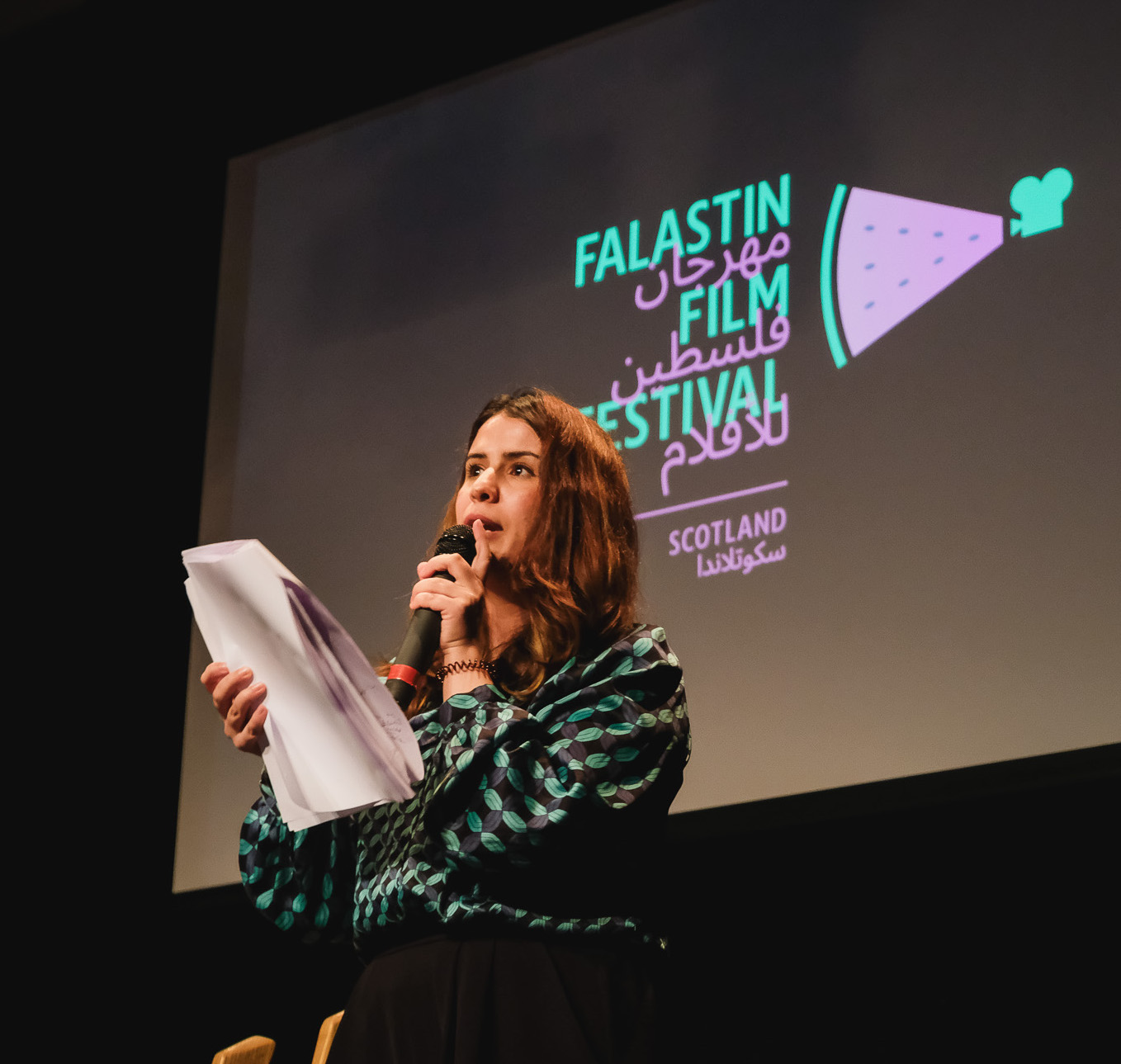 Bayan Haddah reading from notes at a microphone during the Falastin Film Festival 2024 final event, with the festival logo projected behind her.
