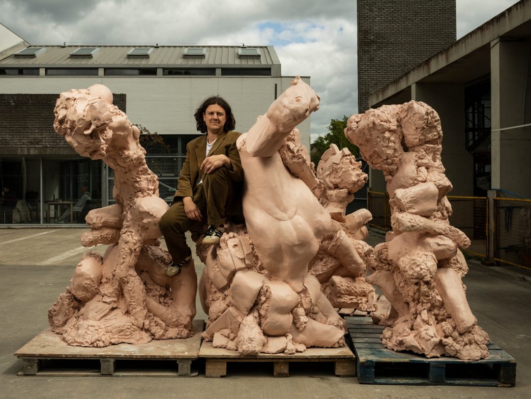 Artist Jamie Fitzpatrick seated atop large, monumental pinkish-brown sculptural works outdoors on wooden pallets.