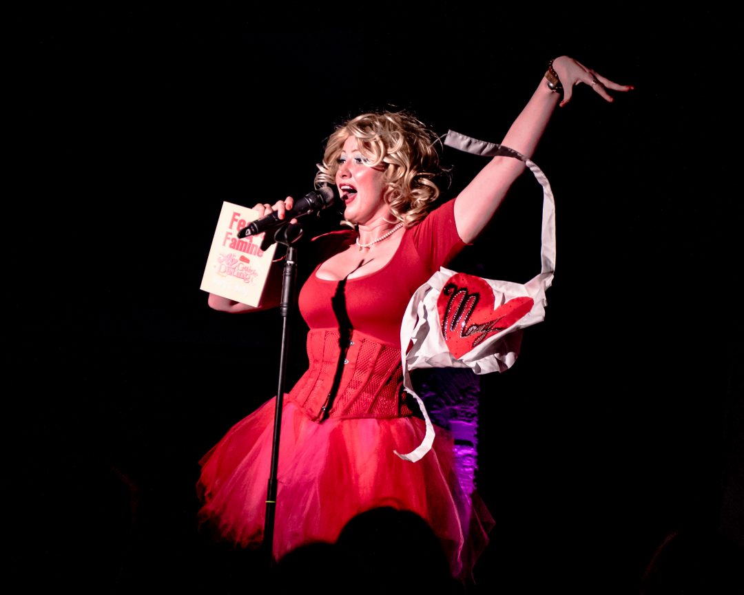 A blond performer in a red dress and corset sings into a microphone on stage, holding a book in one hand while dramatically pulling off a white apron with a red heart.