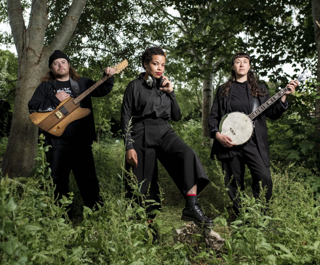 Malaika Kegode and folk-prog duo Jakabol standing outdoors among lush green trees and foliage, wearing dark clothing. Kegode stands centrally with headphones around her neck, while the two band members pose on either side holding electric guitars and a banjo. Photo credit: Paul Blakemere