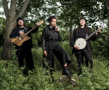 Malaika Kegode and folk-prog duo Jakabol standing outdoors among lush green trees and foliage, wearing dark clothing. Kegode stands centrally with headphones around her neck, while the two band members pose on either side holding electric guitars and a banjo. Photo credit: Paul Blakemere