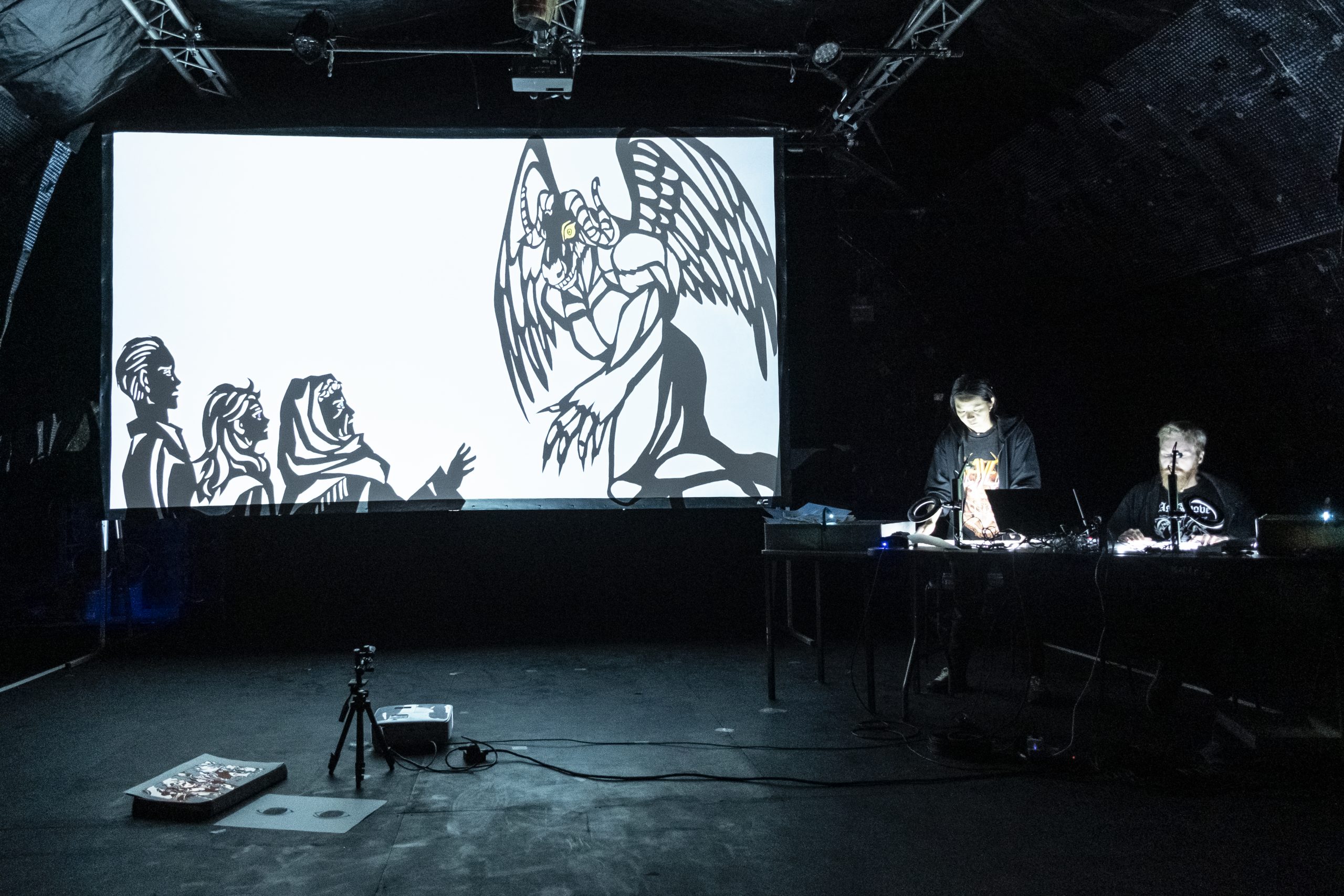 Two puppeteers standing at tables operating shadow puppets, with a large projected screen showing silhouetted characters facing a winged monster.