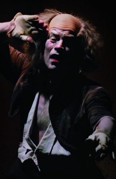 A theatrical performer with white face makeup and a bald cap, holding a gloved hand up near their forehead under dramatic stage lighting.