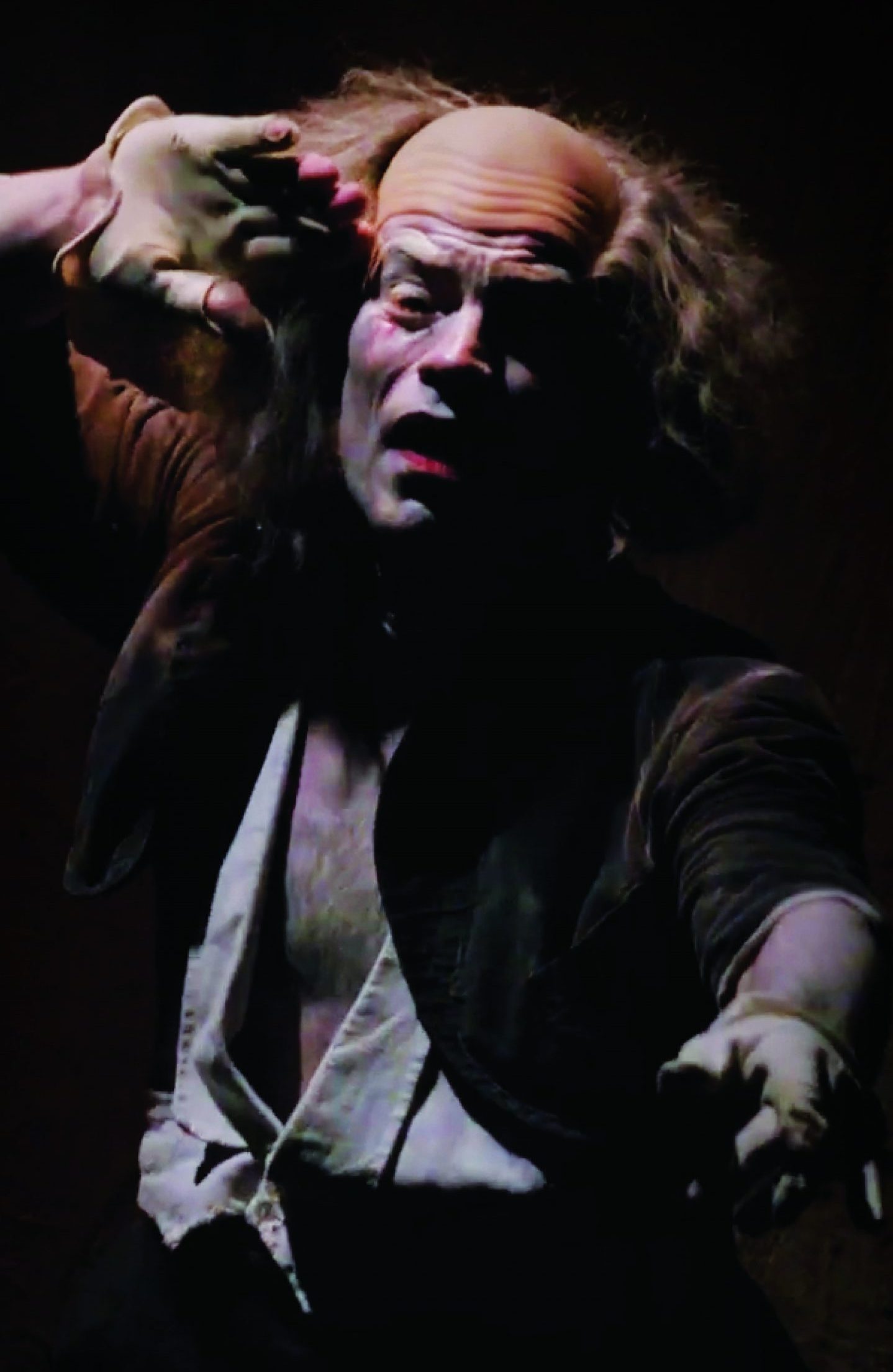 A theatrical performer with white face makeup and a bald cap, holding a gloved hand up near their forehead under dramatic stage lighting.