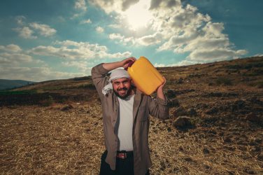 Alaa Shehada smiling while carrying a bright yellow container on his shoulder outdoors in a dry landscape under a cloudy sky.