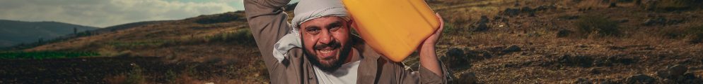 Alaa Shehada smiling while carrying a bright yellow container on his shoulder outdoors in a dry landscape under a cloudy sky.