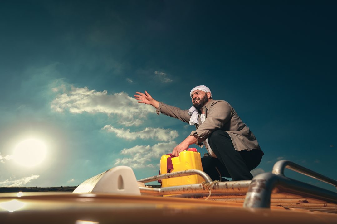 Alaa Shehada crouching on the roof rack of a vehicle next to a yellow container, gesturing with his hand against a bright sky.