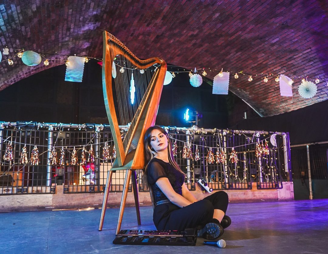 Musician Dara Dubh sitting on stage leaning against a standing harp beneath fairy lights