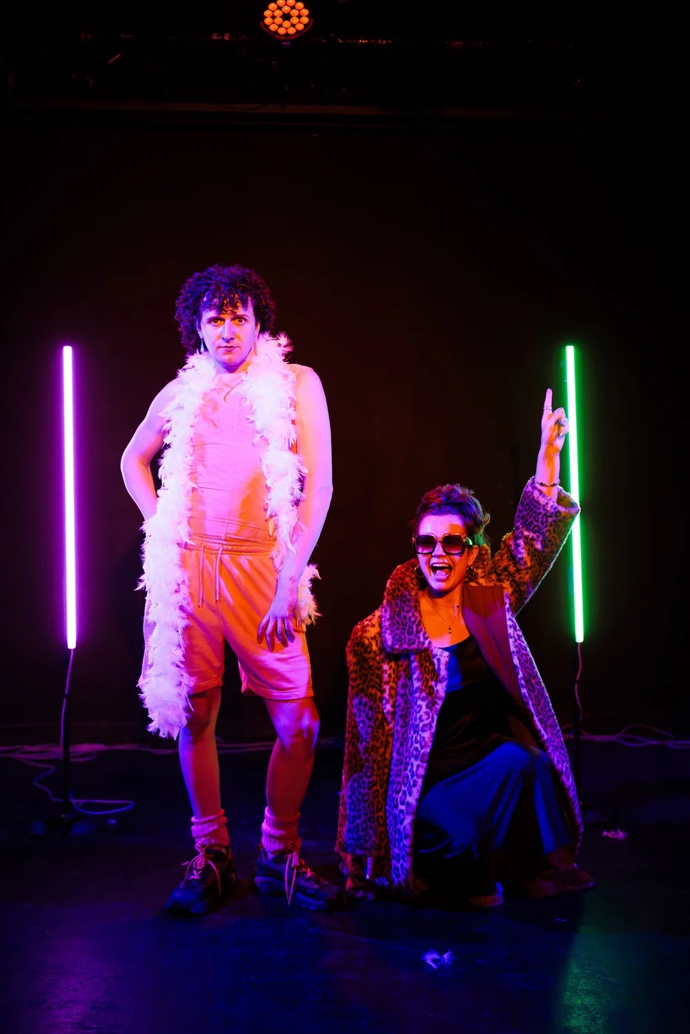 Two performers on stage in front of neon purple and green vertical lights for Desperate Wee Gay Boy; one stands wearing a pink outfit and a white feather boa, while the other crouches in a leopard-print coat and sunglasses.
