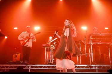 Talulah Ruby singing centre-stage with a microphone during Maribou State's live set, flanked by a guitarist and keyboardist under bright red stage lighting.