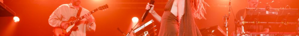 Talulah Ruby singing centre-stage with a microphone during Maribou State's live set, flanked by a guitarist and keyboardist under bright red stage lighting.