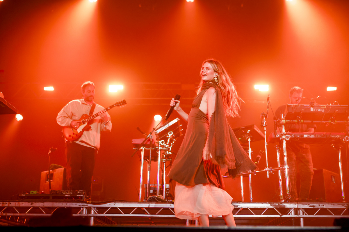 Talulah Ruby singing centre-stage with a microphone during Maribou State's live set, flanked by a guitarist and keyboardist under bright red stage lighting.