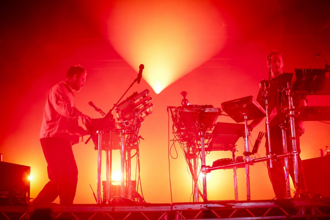 Two members of Maribou State performing live on stage behind electronic instrument rigs under dramatic red lighting.