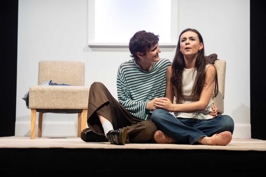 Two actors sitting on the floor side by side holding hands during a performance of For Dolores by Fishamble.