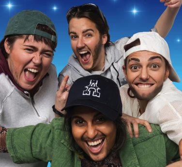 Four performers dressed in casual streetwear, caps, and fake facial hair posing together around a chair against a white background.