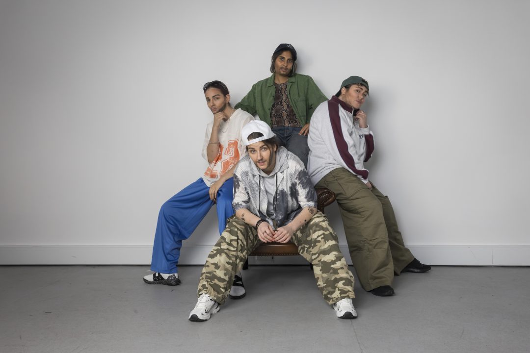 Four performers dressed in casual streetwear, caps, and fake facial hair posing together around a chair against a white background.