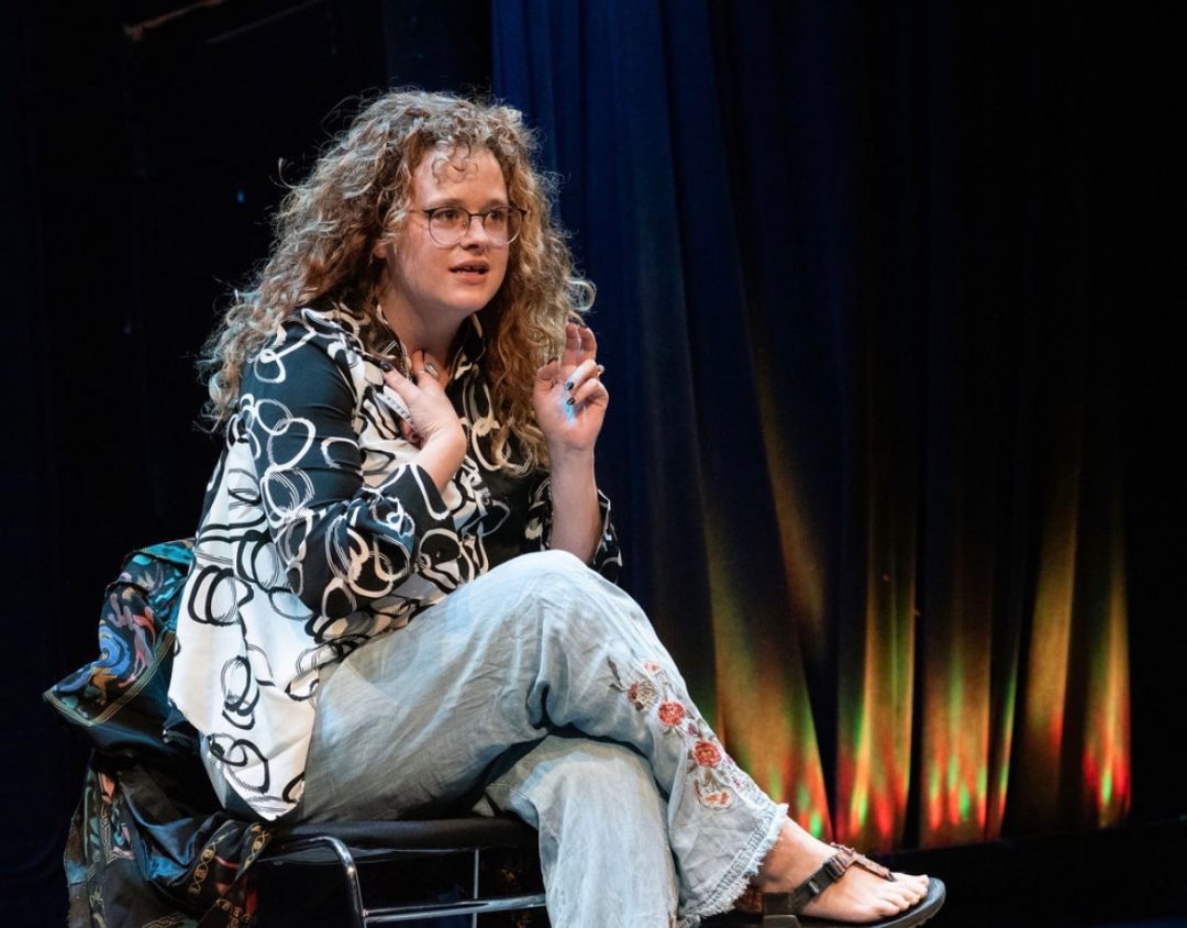 Sara Malinowski on stage with curly hair, glasses, a patterned black and white shirt, and embroidered jeans, seated with her hands raised.