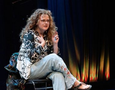 Sara Malinowski on stage with curly hair, glasses, a patterned black and white shirt, and embroidered jeans, seated with her hands raised.