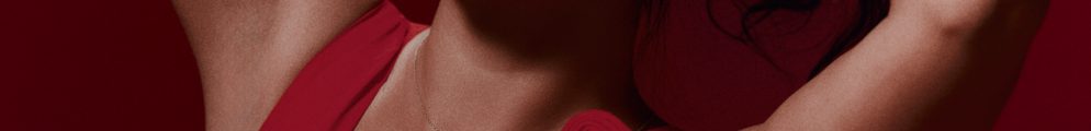 Studio portrait of comedian Jenny Gorelick against a deep red background, wearing a vibrant red dress with a rose detail, looking directly at the camera with hands behind her head, promoting her show Jenny Gorelick: SORRY.