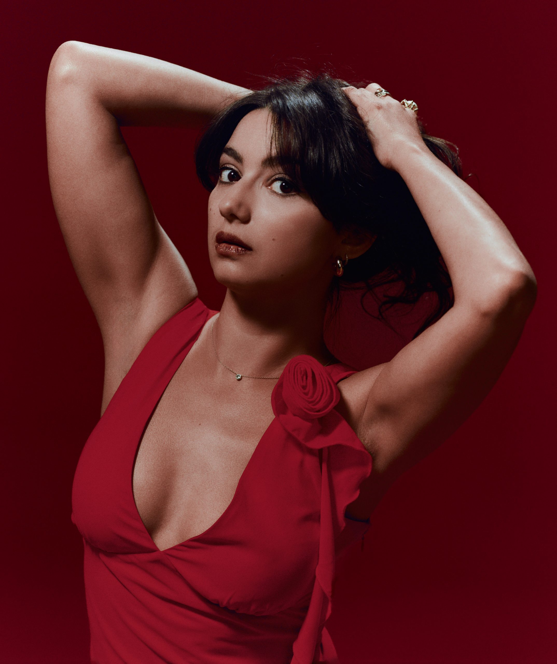 Studio portrait of comedian Jenny Gorelick against a deep red background, wearing a vibrant red dress with a rose detail, looking directly at the camera with hands behind her head, promoting her show Jenny Gorelick: SORRY.